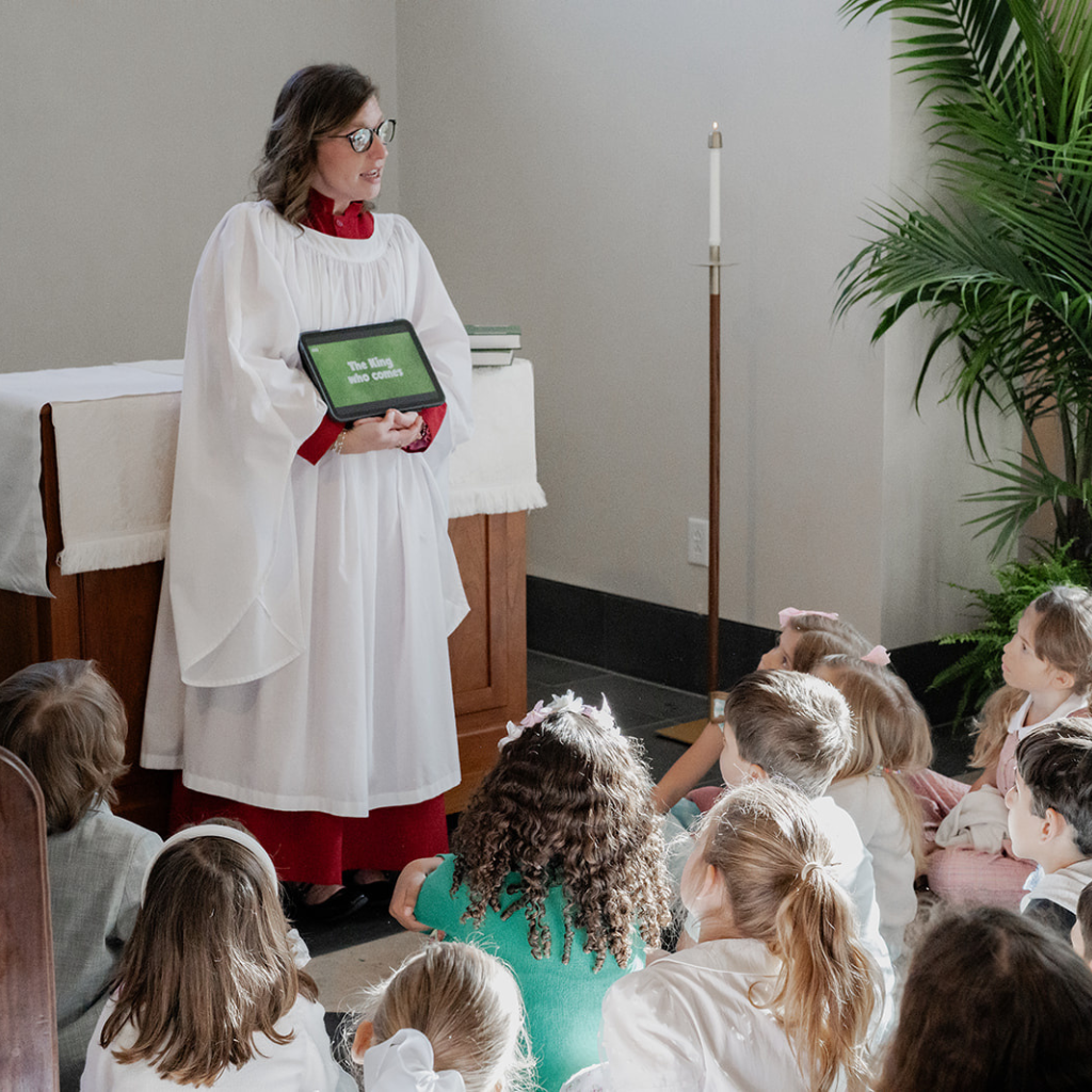 A woman in white vestments holds a tablet reading &lsquo;The King Who Comes!&rsquo; while speaking to seated children.
